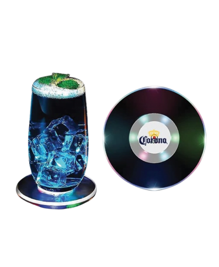 A glass of blue drink with ice and mint sits on a round, multicolored LED Coaster next to another LED Coaster featuring the Corona logo.